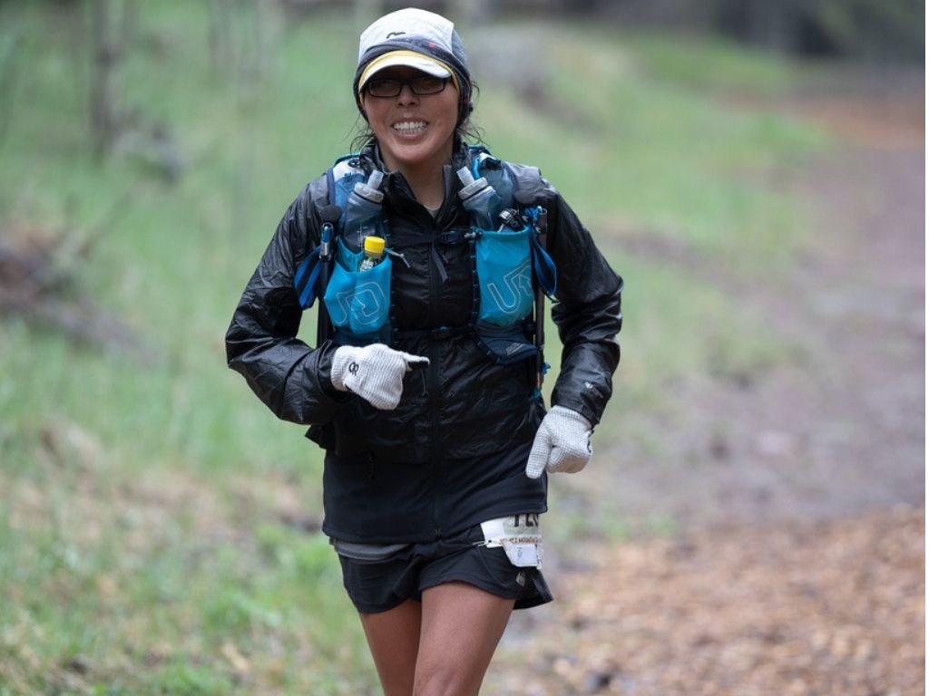 A runner in a race smiling at the camera with a hydration pack, gloves, jacket, and hat on with grass and a trail behind her